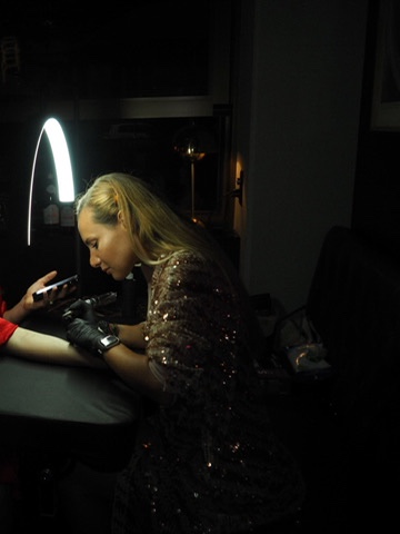 Mina tattooing at an event, in a sequined dress, lit by a ring light